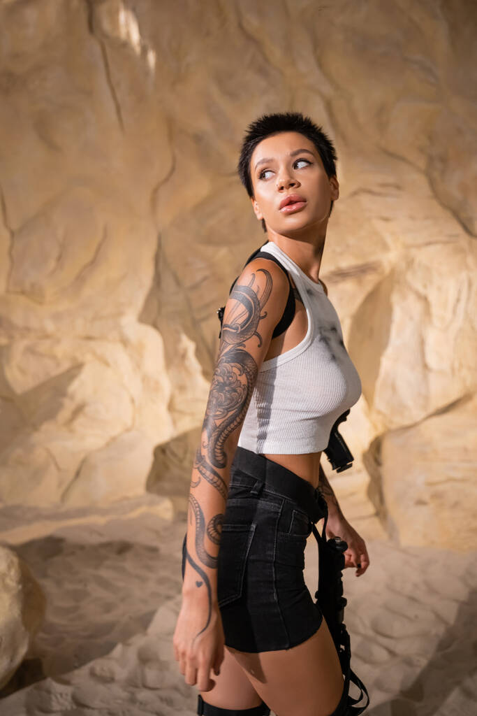 aseistettu ja tatuoitu arkeologi seksikäs asu katselee pois kävellessään luolassa - Valokuva, kuva