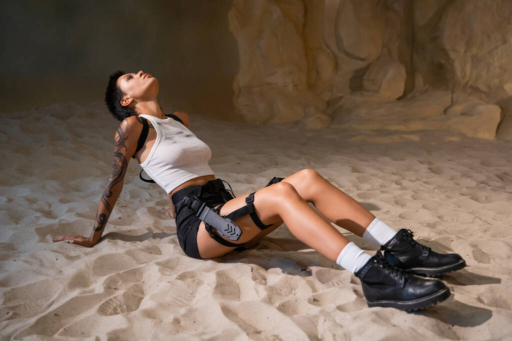 full length of tattooed young woman in dirty sexy clothes sitting on sand - Photo, Image