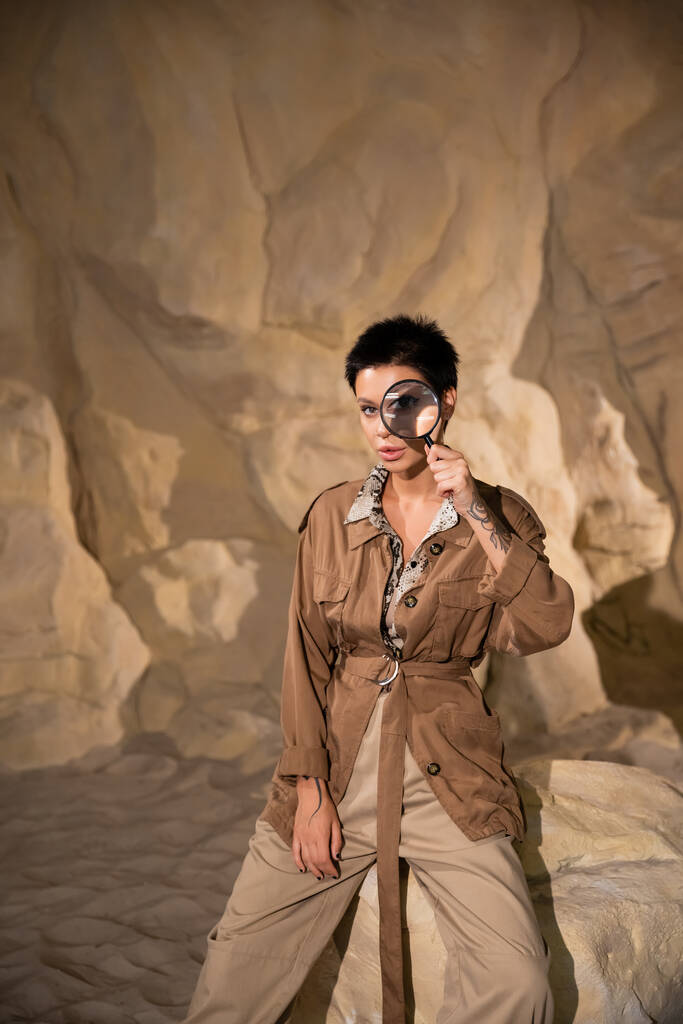 Junge Archäologin mit Tätowierung hält Lupe in der Nähe des Gesichts in Höhle - Foto, Bild