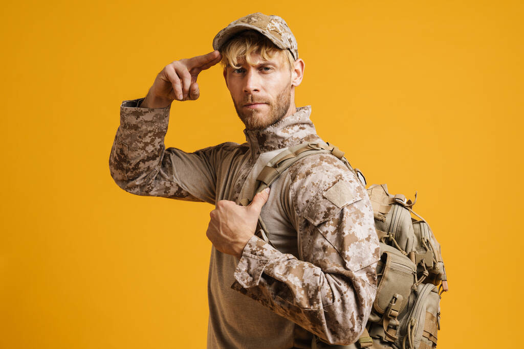 Militar blanco vistiendo uniforme mirando a la cámara y saludando aislado sobre fondo amarillo - Foto, imagen