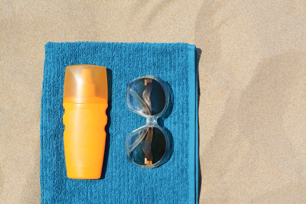 Zachte blauwe strandhanddoek met flesje zonnebrandcrème en zonnebril op zand, plat gelegd. Ruimte voor tekst - Foto, afbeelding