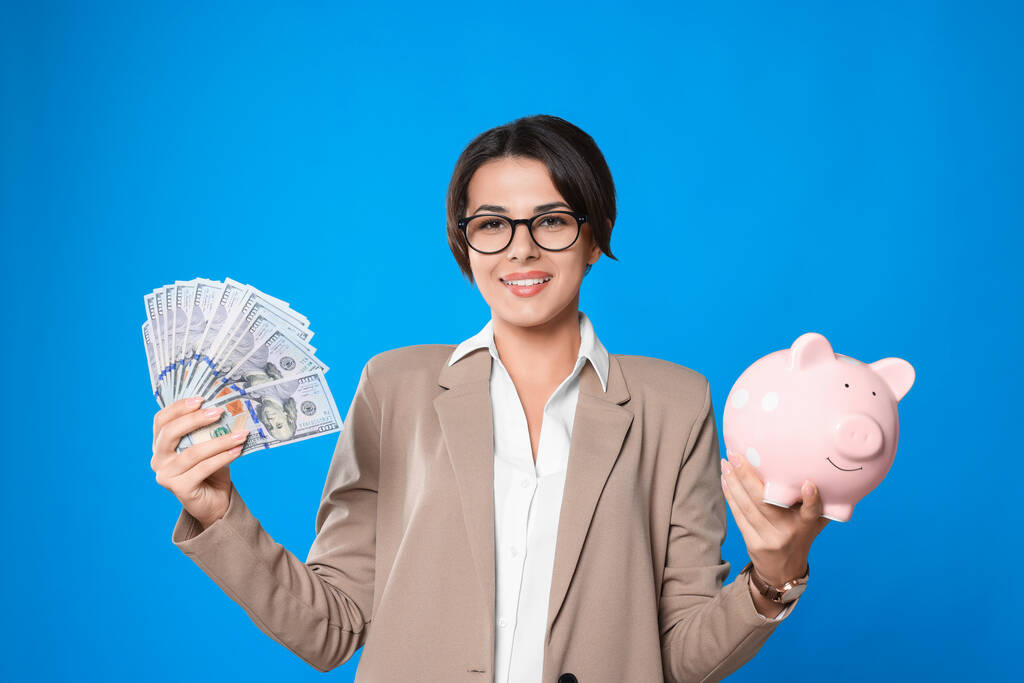 Mujer joven en gafas con dinero y alcancía sobre fondo azul claro - Foto, Imagen