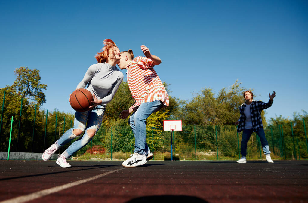 Группа подростков, студенты играют в уличный баскетбол на баскетбольной площадке под открытым небом в весенний солнечный день. Спорт, досуг, хобби, команда, дружба. Мальчики и девочки проводят время вместе. - Фото, изображение