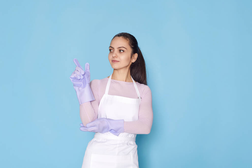 piękna kobieta w fioletowych gumowych rękawiczkach i fartuchu trzymając palec wskazujący z nowym pomysłem na niebieskim tle. sprzątanie - Zdjęcie, obraz