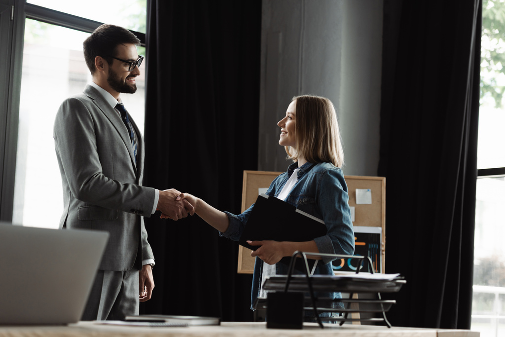 Улыбающаяся женщина держит резюме и пожимает руку бизнесмену во время собеседования в офисе  - Фото, изображение