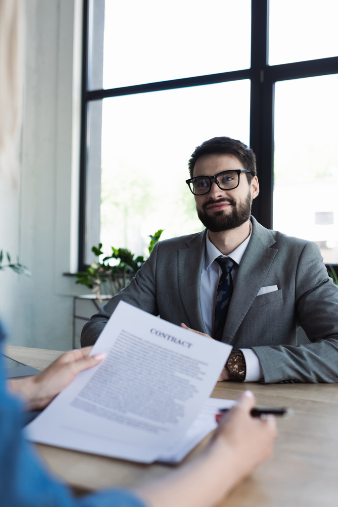 Smiling businessman looking at blurred job seeker with contract in office  - Photo, Image