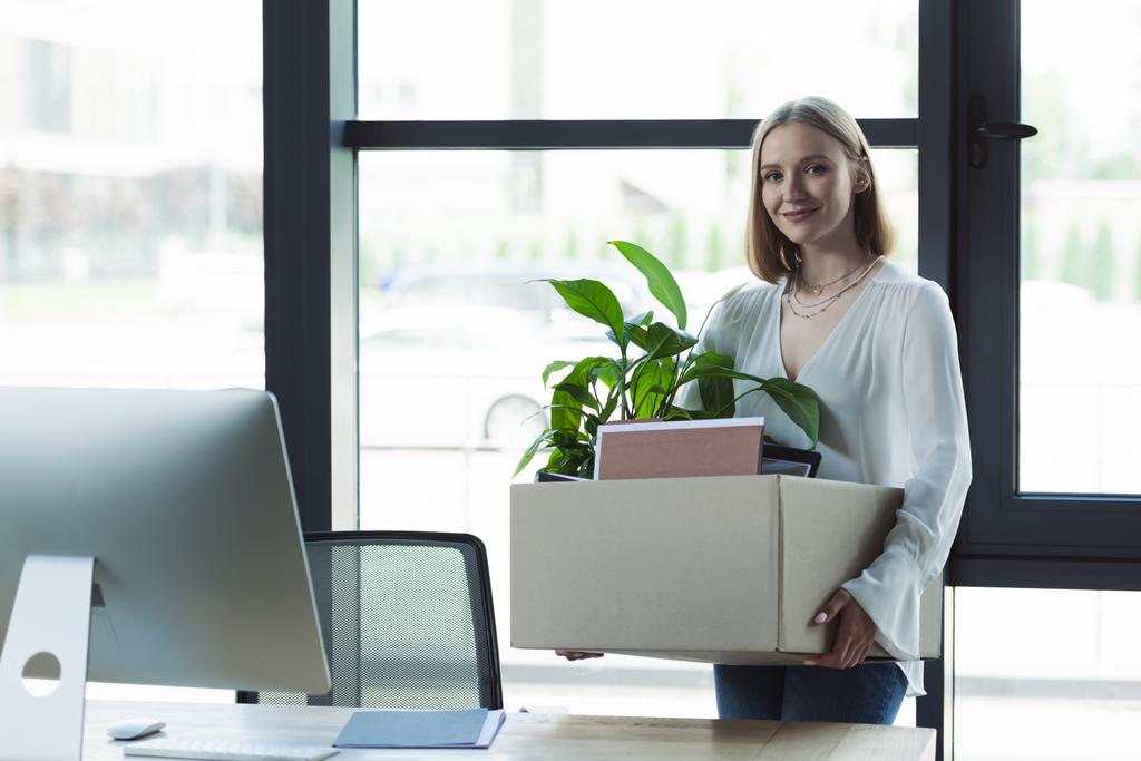 Glimlachende stagiaire met kartonnen doos en kijkend naar camera in de buurt van computer op kantoor  - Foto, afbeelding