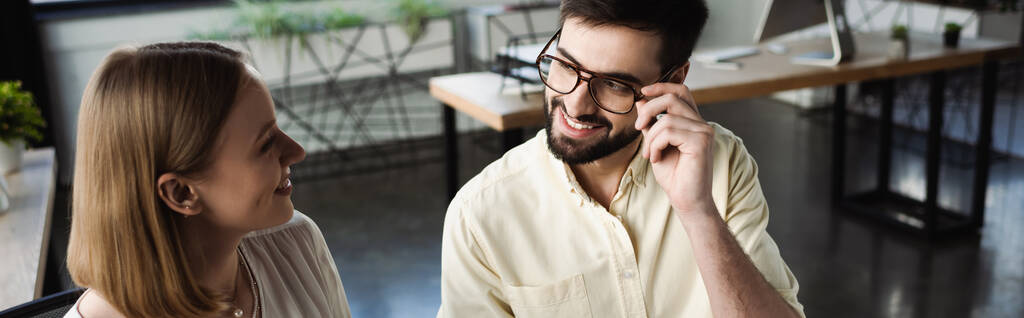 Gestionnaire souriant touchant des lunettes tout en parlant à un stagiaire au bureau, bannière  - Photo, image