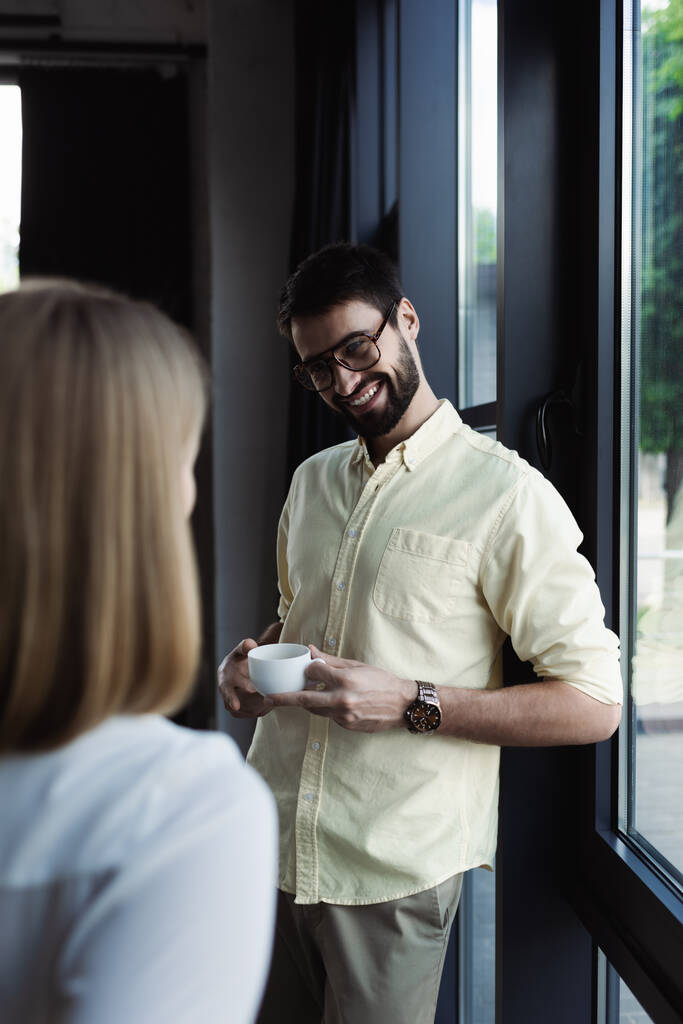 Un hombre de negocios sonriente sosteniendo una taza de café cerca de un interno borroso en la oficina  - Foto, Imagen