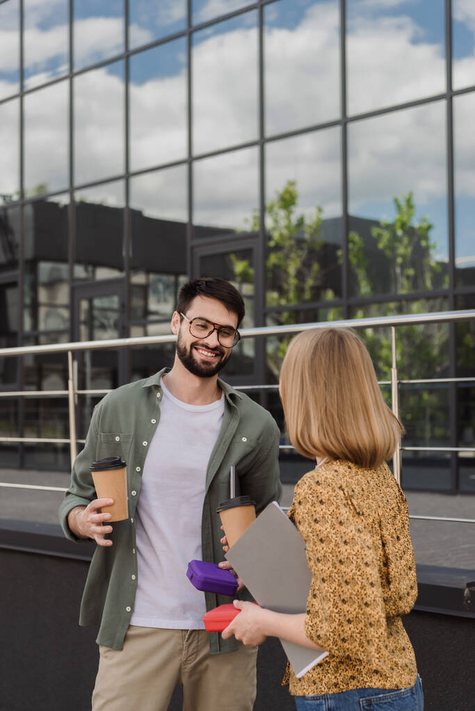 Lächelnder Geschäftsmann mit Lunchbox und Coffee to go, während er mit Kollegen mit Papiermappe im Freien spricht  - Foto, Bild