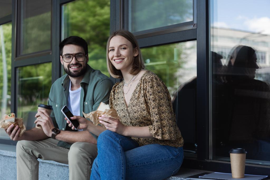 Des hommes d'affaires souriants tenant des sandwichs et un smartphone tout en regardant la caméra près du bâtiment à l'extérieur  - Photo, image