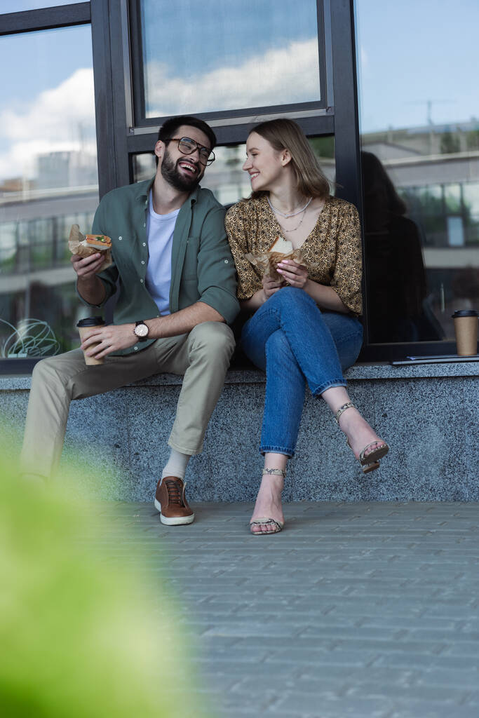 Ενθουσιασμένοι επιχειρηματίες κρατώντας σάντουιτς και καφέ, ενώ μιλάμε κοντά κτίριο σε αστικό δρόμο  - Φωτογραφία, εικόνα