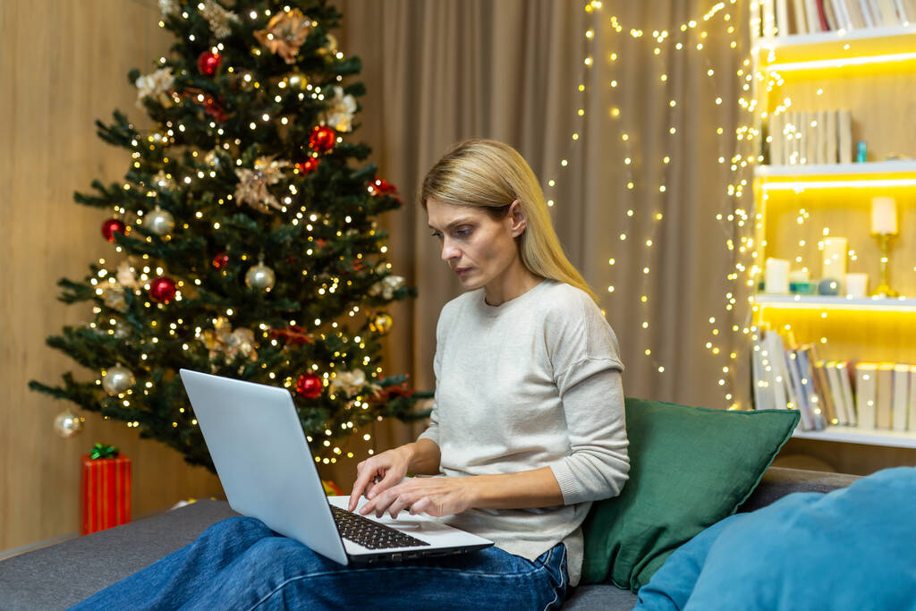 Mujer sola en casa para Navidad, sentado en el sofá y trabajando con el ordenador portátil de forma remota durante las vacaciones de año nuevo, la mujer de negocios pensando y centrado en serio. - Foto, Imagen