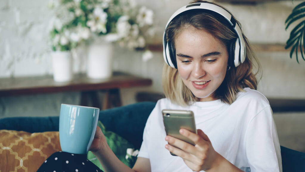 Šťastná mladá dáma používá smartphone online chatování a poslech hudby s bezdrátovými sluchátky, která seděla na pohovce v moderním bytě. Koncept technologie, lidé a radost. - Fotografie, Obrázek