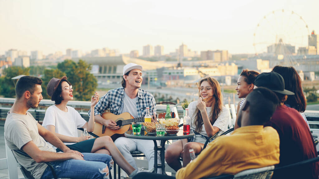 Joyful male musician is playing the guitar with his friends singing and laughing sitting on rooftop at table. Entertainment, musical instruments and leisure concept. - Photo, Image