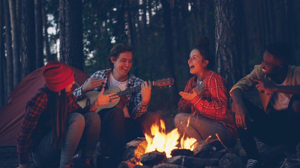 jonge man toerist speelt gitaar terwijl zijn vrienden zingen en lachen zitten rond vuur in het bos in de avond genieten van de natuur en goed gezelschap. - Foto, afbeelding