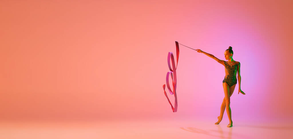 Studio photo de jeune fille charmante, entraînement rythmique gymnaste avec des équipements sportifs isolés sur fond rose dans un filtre lumineux au néon. Danse, musique, émotions, défis, motivation et publicité - Photo, image