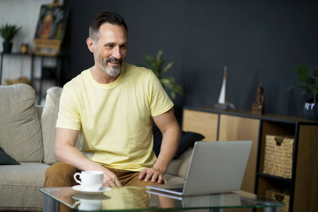 Laptopot használ, amelynek online konferencia az ügyfelek középkorú férfi ül a kanapén, kanapé mellett üveg asztal egy csésze kávét. Érett jóképű szabadúszó IT férfi dolgozik számítógép otthon.  - Fotó, kép