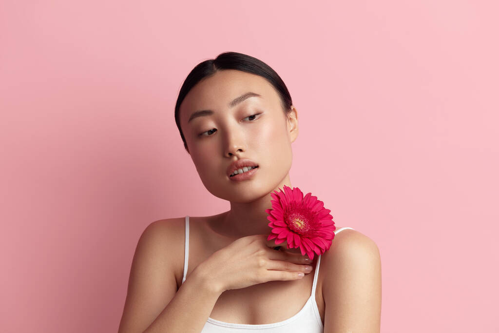 Hautpflegerin posiert mit roter Blume. Asiatisches Modell mit Blume vor der Kamera Weiblich mit nacktem Make-up, glatter, hydratisierter und strahlender Haut. Natürliche Schönheit auf rosa Hintergrund  - Foto, Bild