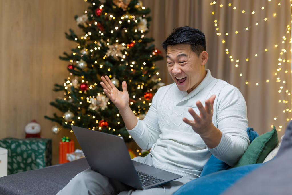 Aasialainen mies lukee iloisia uutisia kannettavalla tietokoneella kotona jouluksi, liikemies työskentelee etänä uudenvuoden lomien aikana istuu sohvalla olohuoneessa, juhlii menestystä.. - Valokuva, kuva