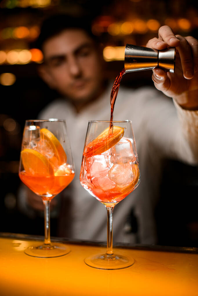 läpinäkyvä lasi jääkuutioilla ja appelsiiniviipaleilla baaritiskillä ja baarimikon käsi kaataa juomaa teräsjiggeristä lasiin - Valokuva, kuva