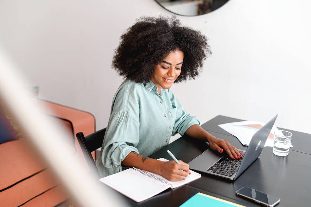 Uśmiechnięta młoda, wielorasowa kobieta pracuje w domu. Niezależna kobieta siedząca przy biurku w biurze przed laptopem, robiąca notatki. Kobieta ucząca się na odległość - Zdjęcie, obraz