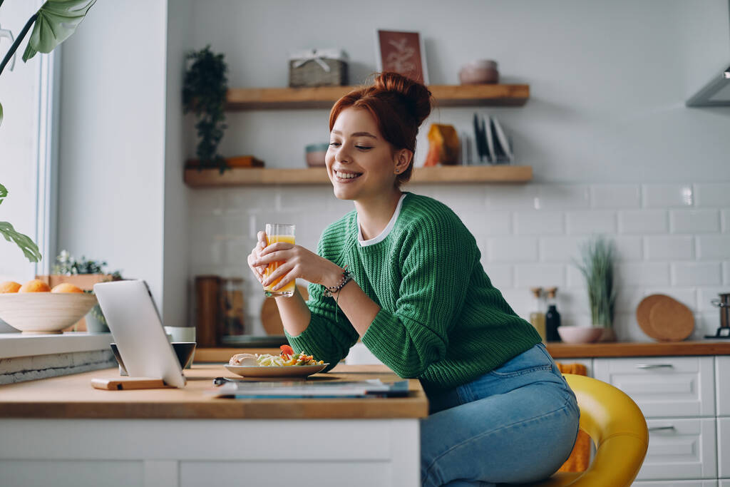 Красивая молодая женщина обедает и смотрит на цифровой планшет, сидя за кухонным столом - Фото, изображение
