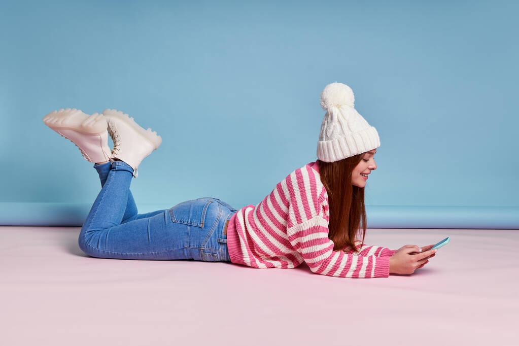 Hermosa mujer en ropa de invierno utilizando el teléfono inteligente y sonriendo mientras está tumbado sobre fondo azul - Foto, imagen