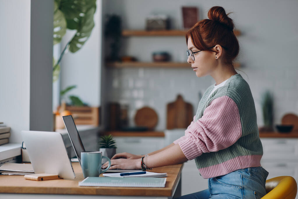 Fiatal vörös hajú nő dolgozik laptop, miközben ül a konyhapulton otthon - Fotó, kép