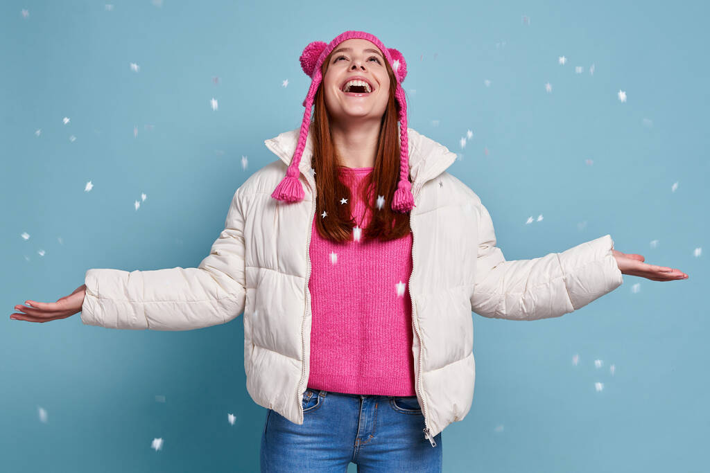 Gelukkig vrouw in winterjas houden armen uitgestrekt terwijl sneeuw valt tegen een blauwe achtergrond - Foto, afbeelding