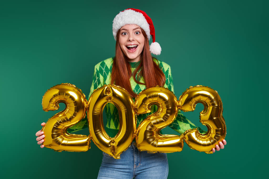 Noel şapkalı mutlu kadın yeşil arka planda altın renkli numaralar tutuyor. - Fotoğraf, Görsel