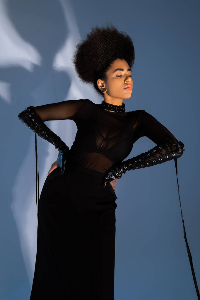 modelo afroamericano de moda en traje negro posando con las manos en las caderas en azul  - Foto, Imagen