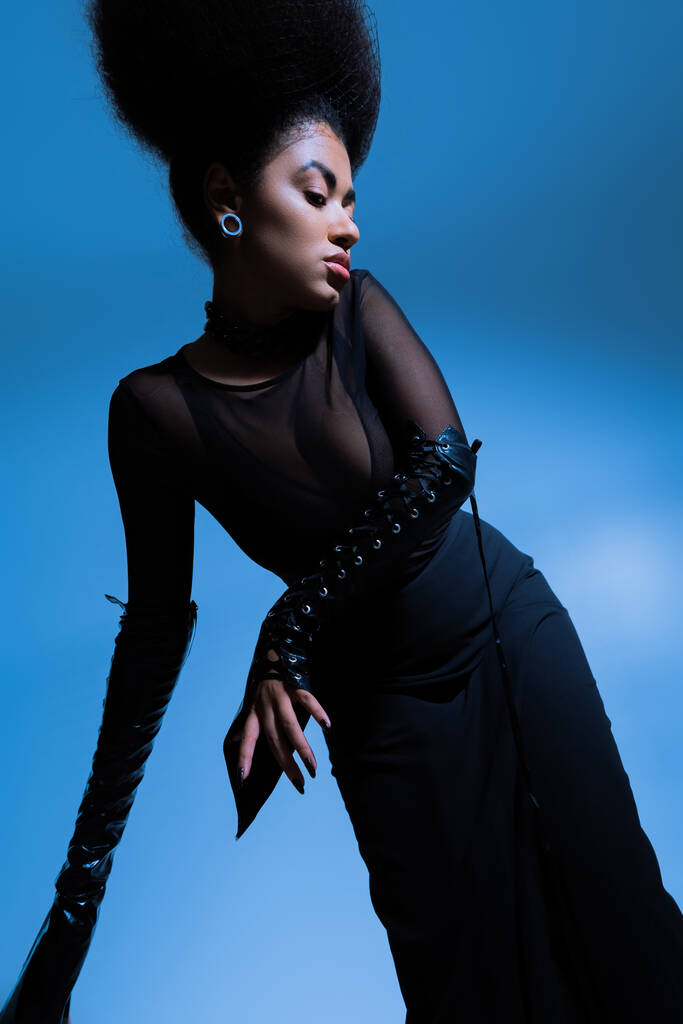 Siyah kıyafetli kıvırcık Afrikalı Amerikan mankeni mavi tarafa bakarken poz veriyor. - Fotoğraf, Görsel