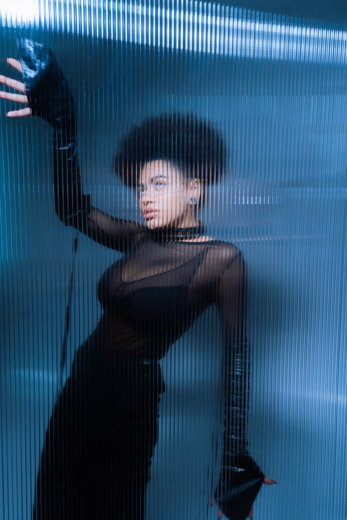 Genç Afro-Amerikalı seksi siyah elbiseli kadın fırfırlı camın arkasında poz veriyor. - Fotoğraf, Görsel
