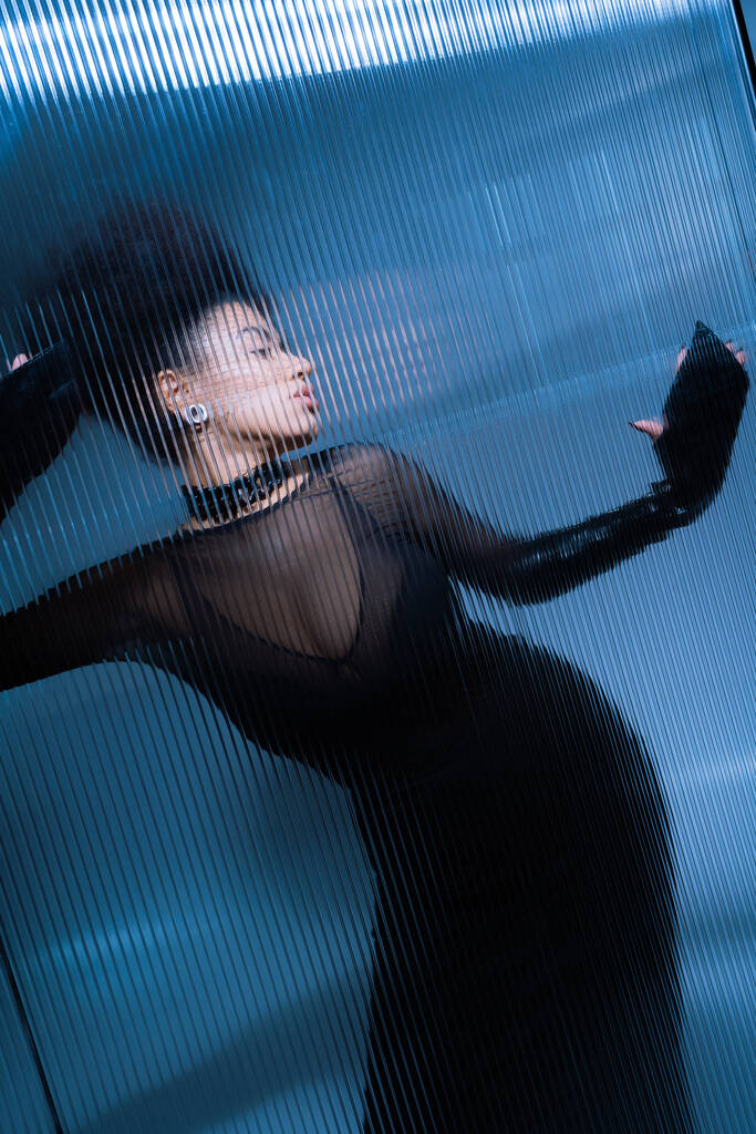 Genç Afro-Amerikan modeli, siyah seksi elbisesiyle dalgalı camların arkasında poz veriyor. - Fotoğraf, Görsel