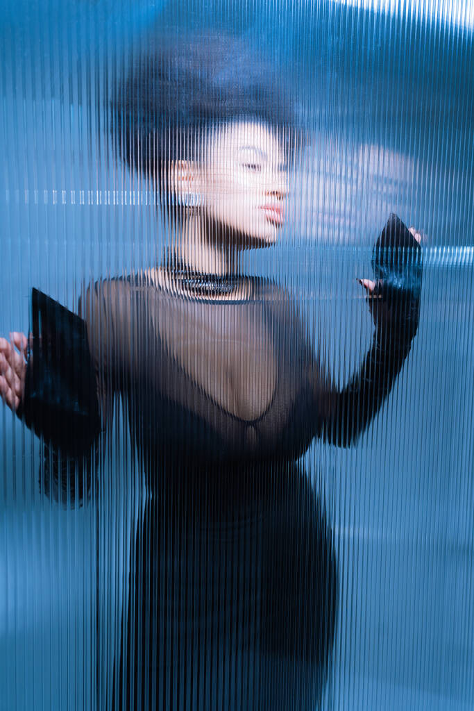 ładny afrykański amerykański model w czarny sexy strój patrząc daleko podczas dotykania szorstkie szkło - Zdjęcie, obraz