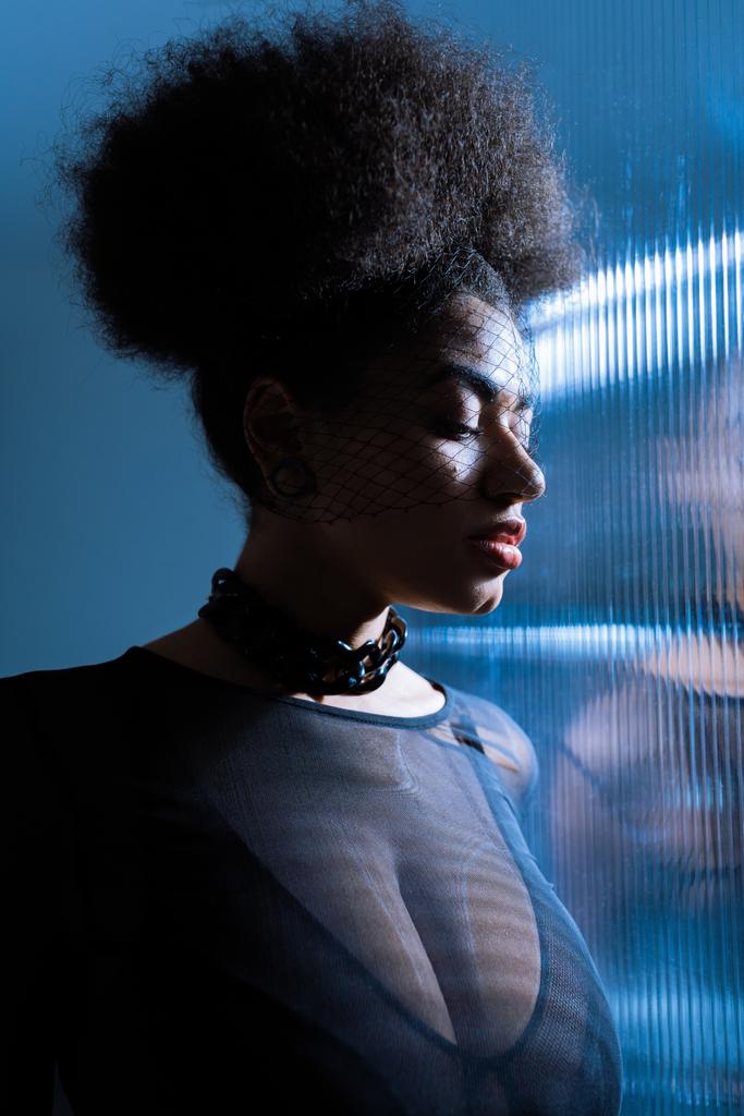 mladá africká americká žena v sexy oblečení a černý závoj v blízkosti čalouněné sklo s modrým osvětlením  - Fotografie, Obrázek