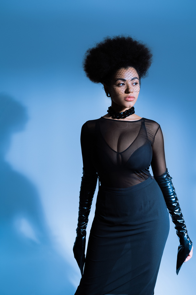 afro-américaine en tenue sexy et voile noir regardant loin tout en posant sur le bleu  - Photo, image