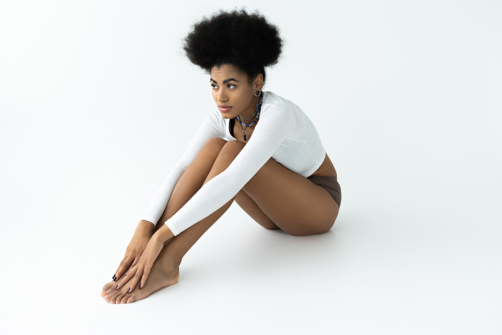 Uzun kollu gömlek ve külotlu beyaz arka planda oturan çıplak ayaklı Afro-Amerikan kadın.  - Fotoğraf, Görsel