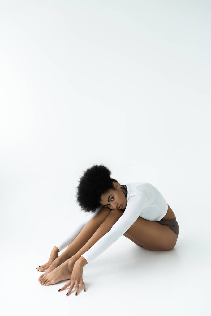 longitud completa de la joven afroamericana mujer en camisa de manga larga y bragas sentado en blanco  - Foto, imagen
