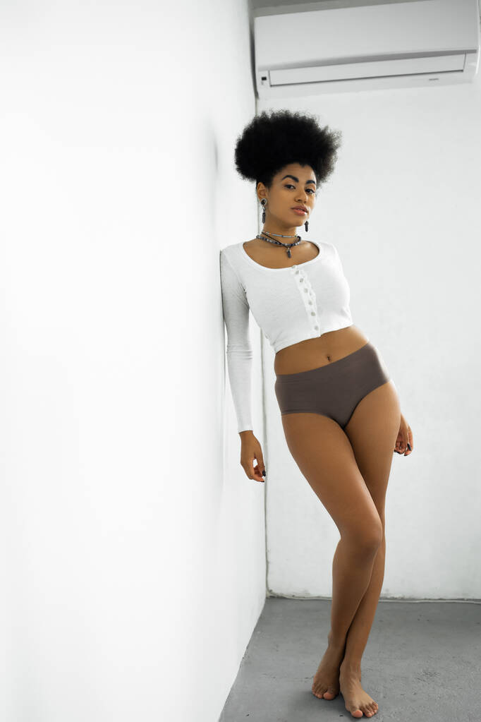 longitud completa de mujer afroamericana descalza en camisa de manga larga y ropa interior de pie cerca de la pared blanca - Foto, imagen
