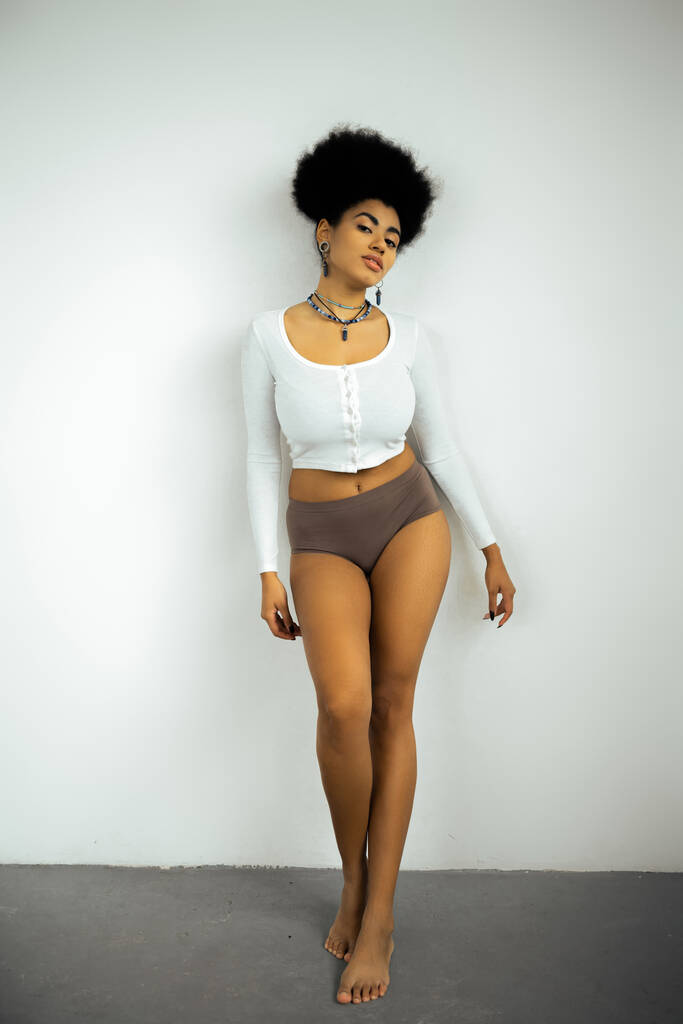 piena lunghezza della donna afroamericana a piedi nudi in camicia a maniche lunghe e mutandine in piedi vicino alla parete bianca  - Foto, immagini