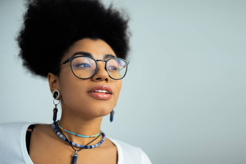 portrait of dreamy african american woman in eyeglasses and accessories isolated on grey  - Photo, Image