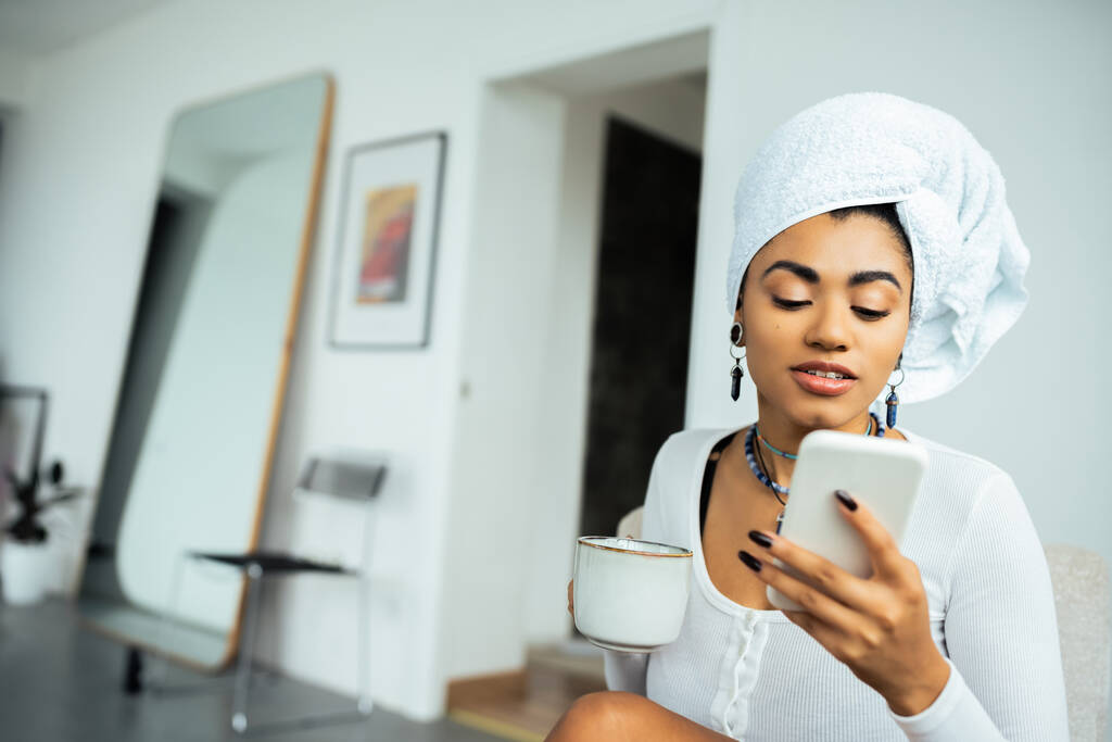 mulher americana africana bonita com toalha na cabeça usando smartphone e segurando xícara de café  - Foto, Imagem