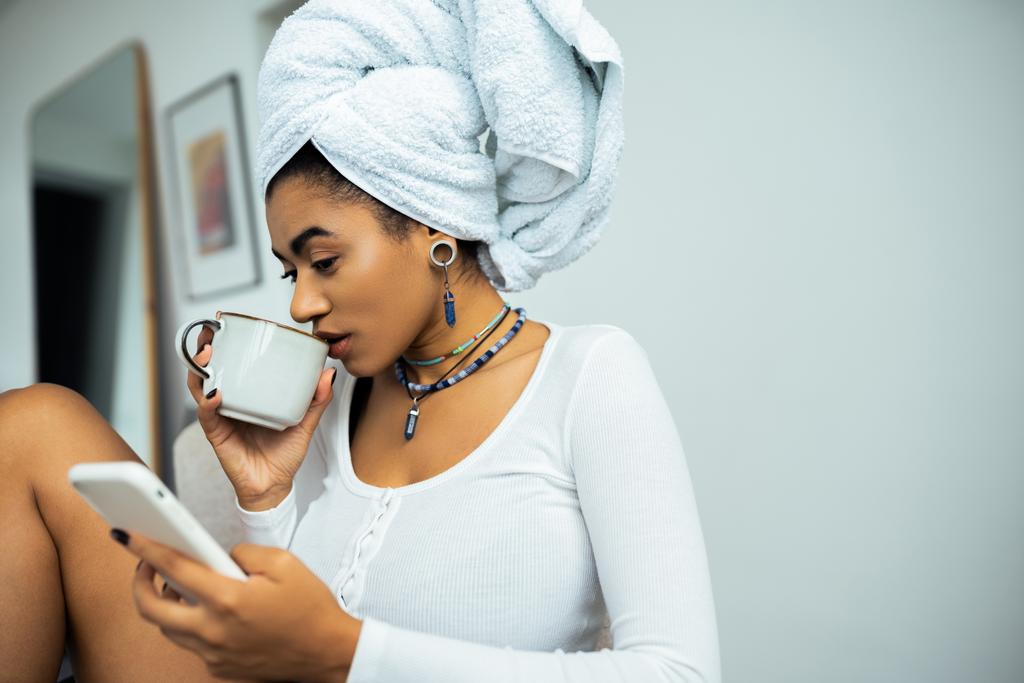 hezká africká americká žena s ručníkem na hlavě pomocí smartphonu a pití kávy  - Fotografie, Obrázek