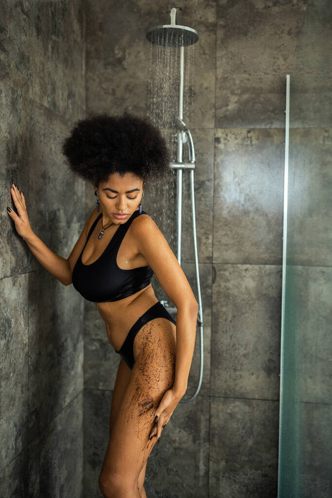 riccio afroamericano donna applicando caffè scrub sotto l'acqua in cabina doccia - Foto, immagini
