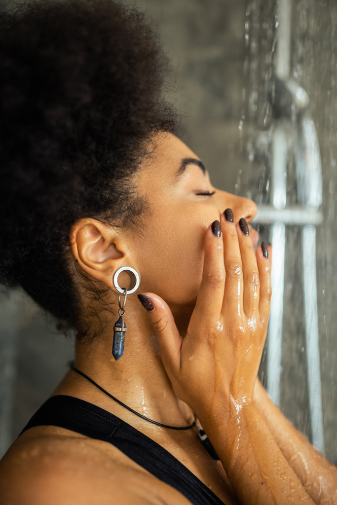 Vista lateral de la mujer afroamericana lavando la cara bajo el agua en la ducha - Foto, imagen