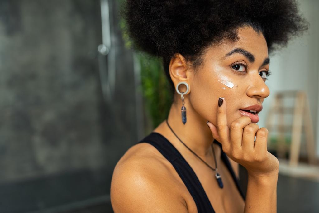 Femme afro-américaine appliquant crème sur le visage dans la salle de bain floue  - Photo, image