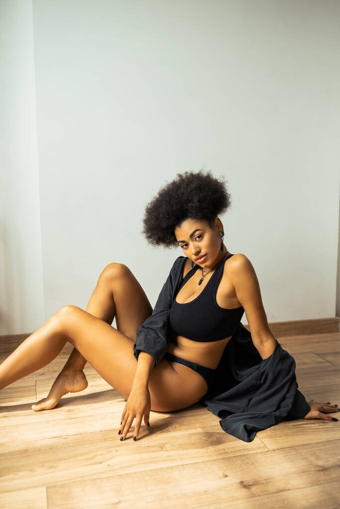 Hübsche afrikanisch-amerikanische Frau in Unterwäsche und Hemd schaut in die Kamera, während sie zu Hause auf dem Boden sitzt  - Foto, Bild