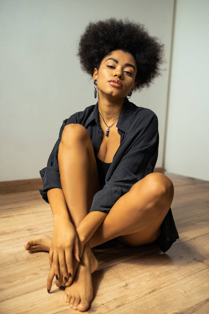 Giovane donna afroamericana in camicia nera guardando la fotocamera mentre seduto sul pavimento  - Foto, immagini